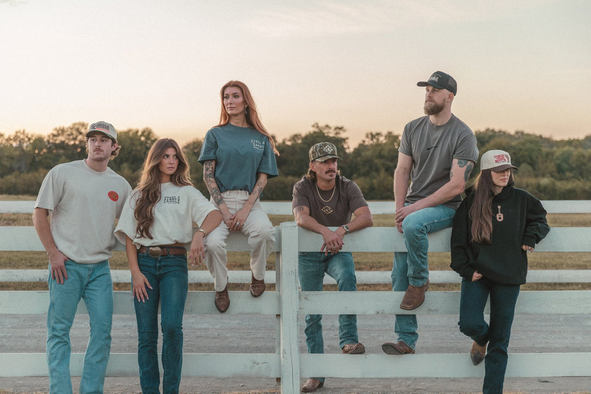 Friends in Steel & Saddle tees and caps sitting on a white ranch fence