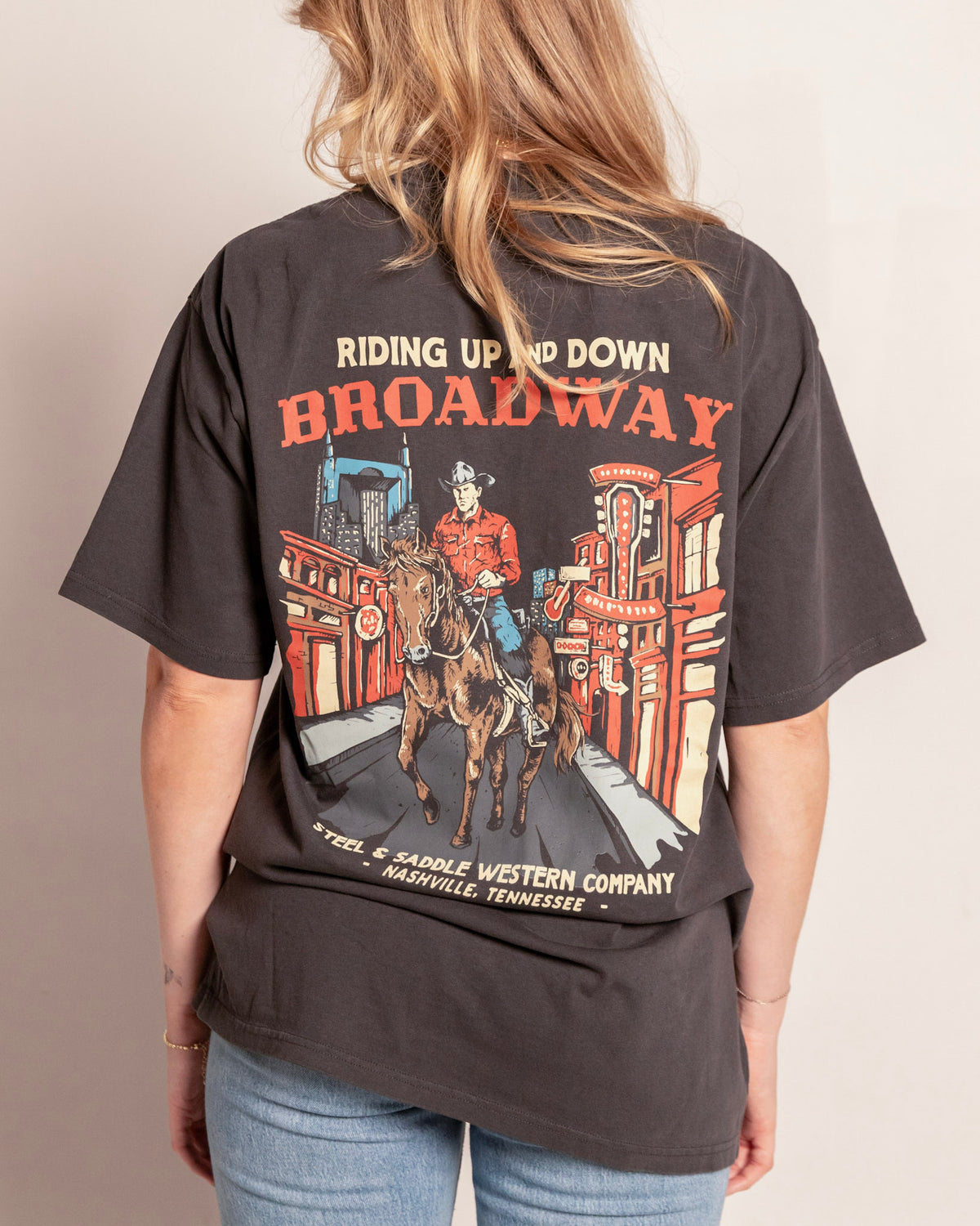 Broadway Tee in faded coal on model, back view with the cowboy riding a horse down neon lit Lower Broadway