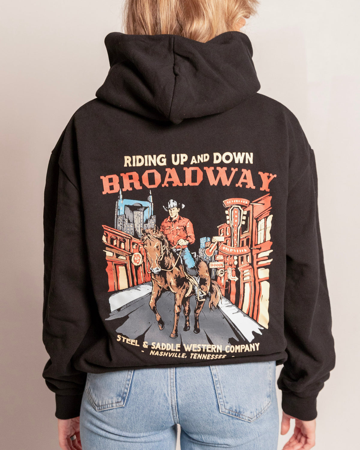 Broadway Hoodie in faded black on model, back view with the cowboy riding a horse down neon lit Lower Broadway