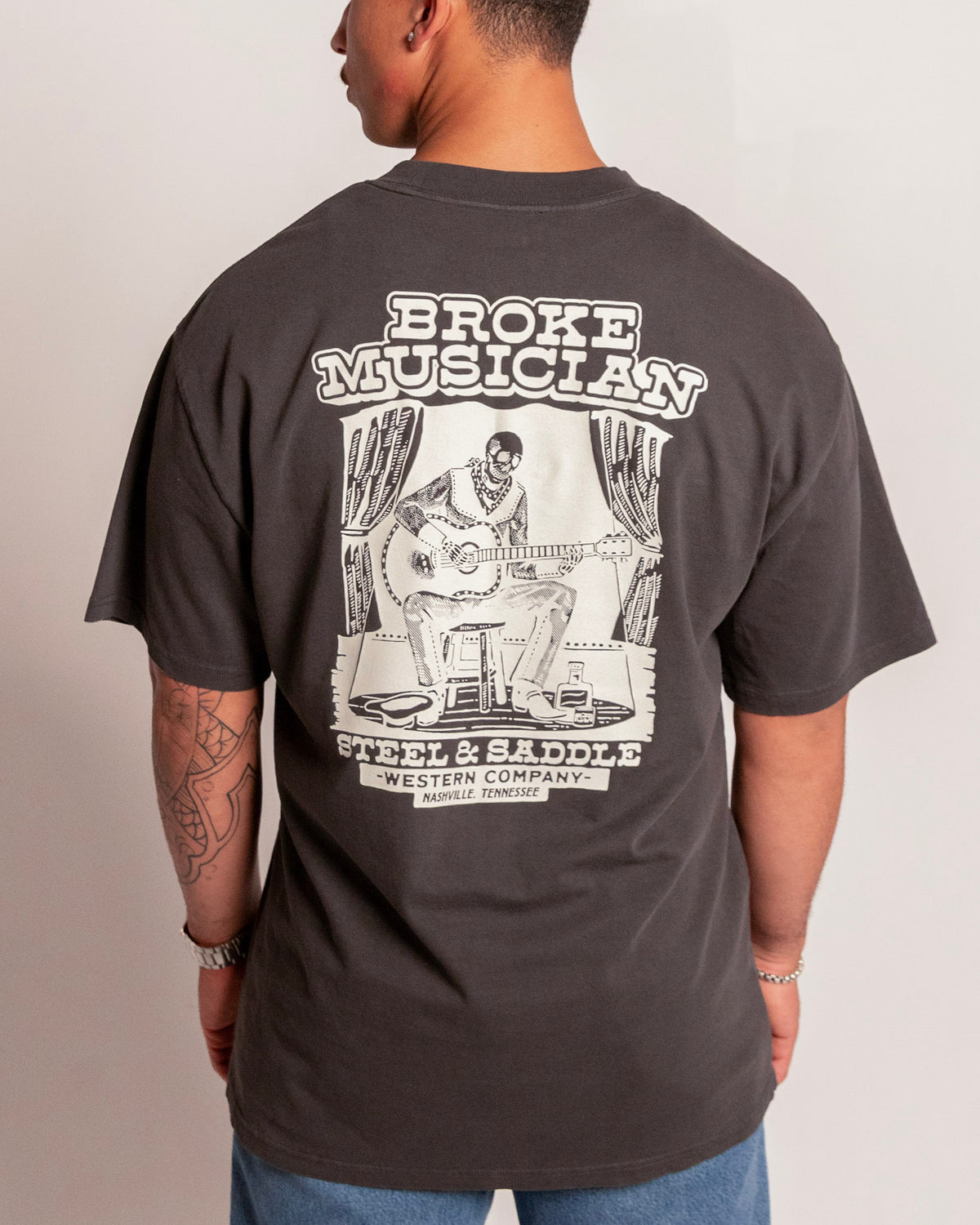Broke Musician Tee in faded coal on model, back view with the skeleton guitarist on a stage stool