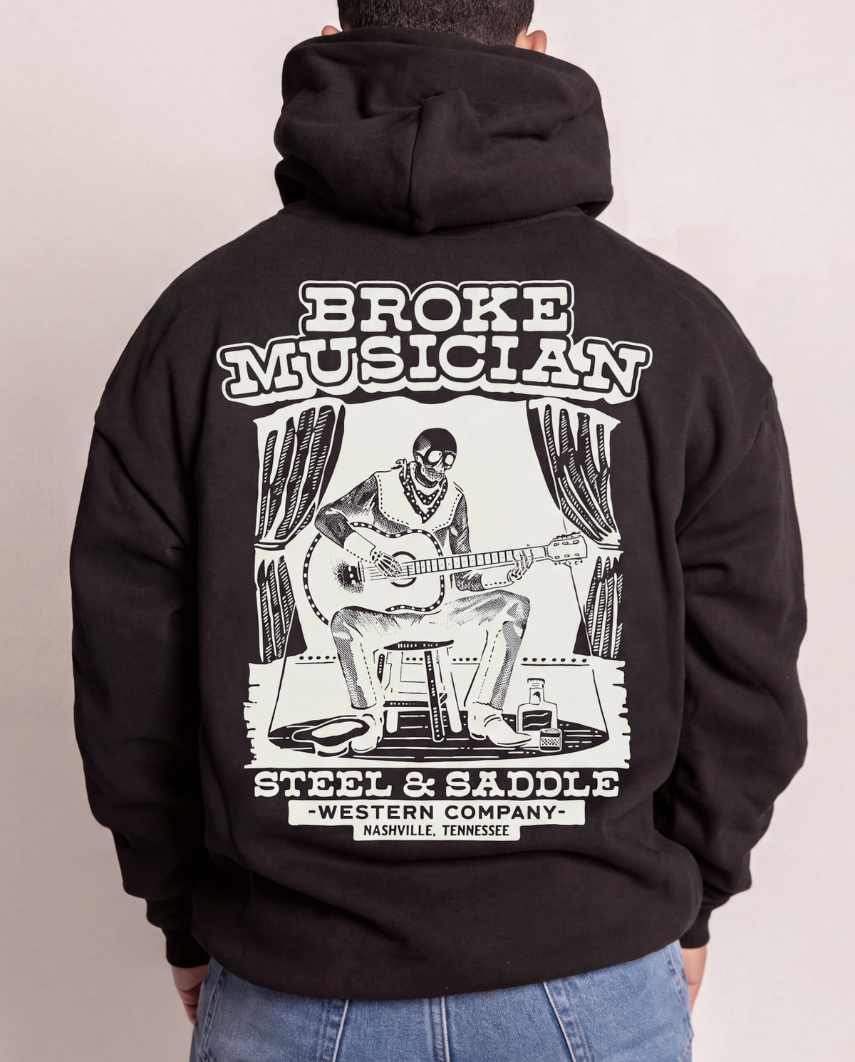 Broke Musician Hoodie in faded black on model, back view with the skeleton guitarist on a stage stool