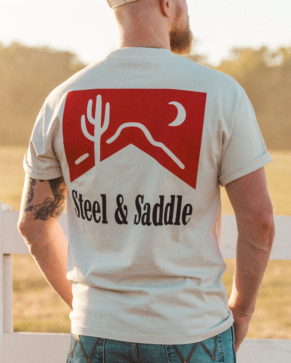 Cowboy Killer Tee in faded bone worn outdoors, back print of a red pack, cactus and moon over Steel & Saddle lettering