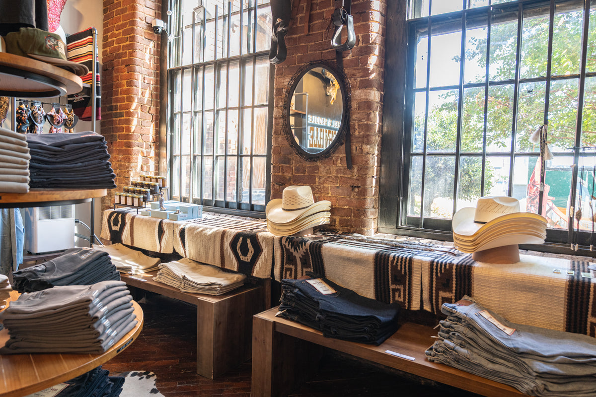 Straw cowboy hats and folded western wear on blanket-draped benches under tall windows at Steel & Saddle, Marathon Village, Nashville