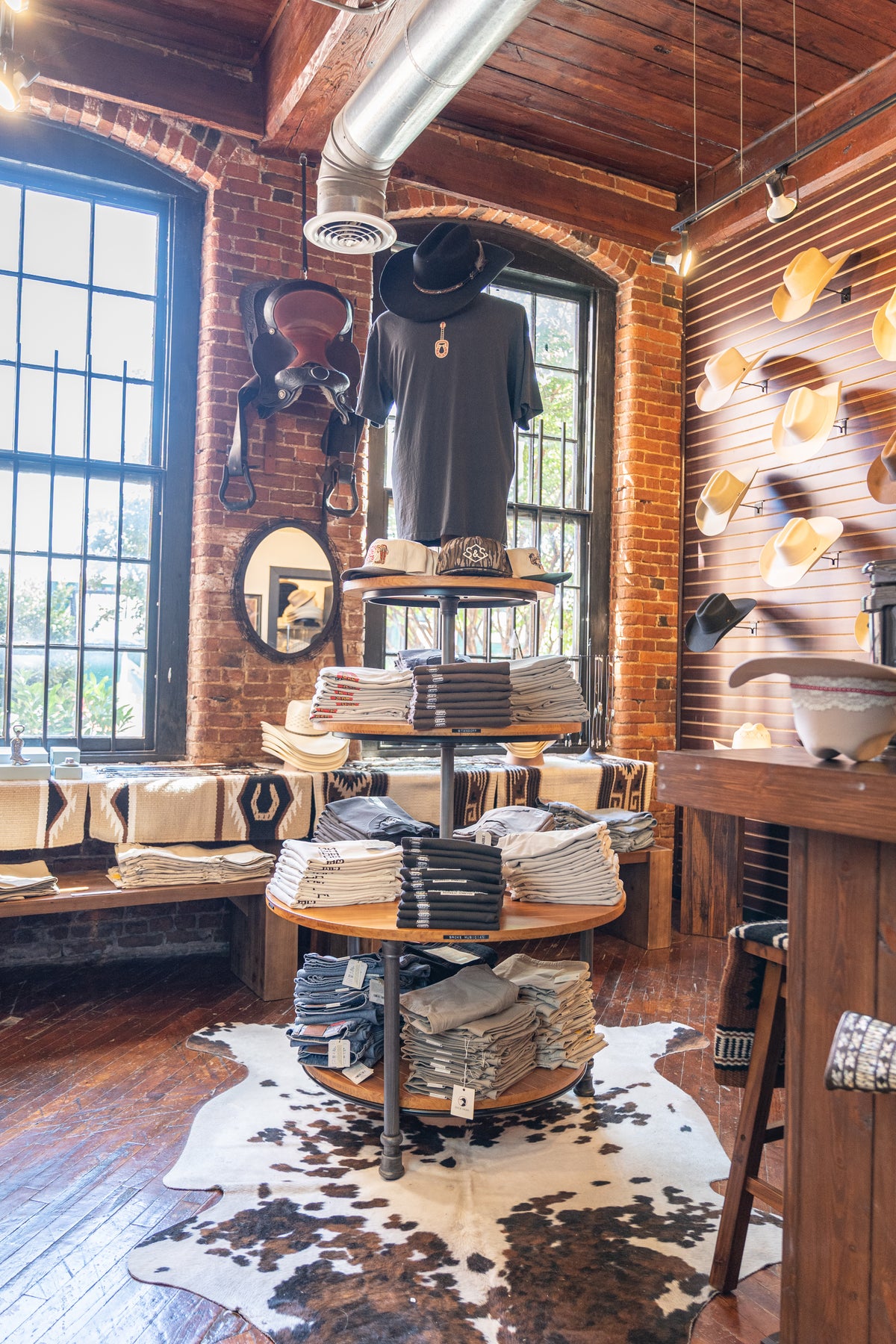 Round wooden display of folded tees on a cowhide rug, with cowboy hats on the brick wall of the Steel & Saddle shop in Nashville