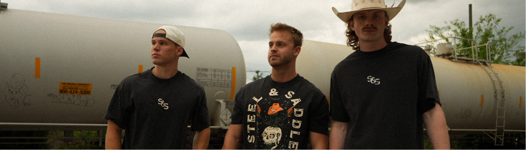 Three men standing in front of a large cylindrical tank with a cloudy sky in the background.