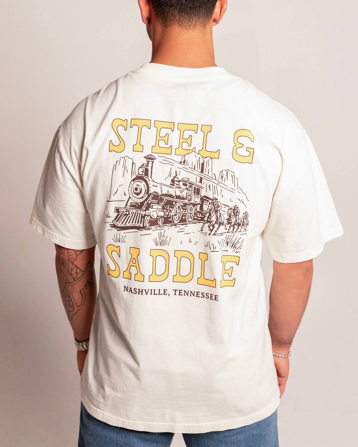 Holdup Tee in faded ecru on model, back view with the riders chasing a steam train between giant Steel & Saddle lettering