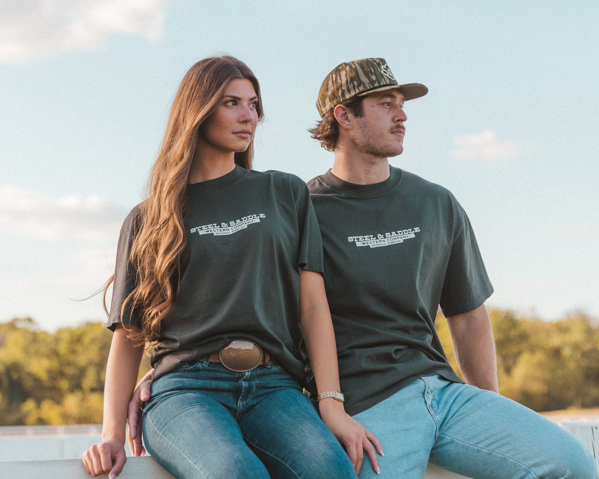 Couple in Steel & Saddle tees sitting together on a white ranch fence