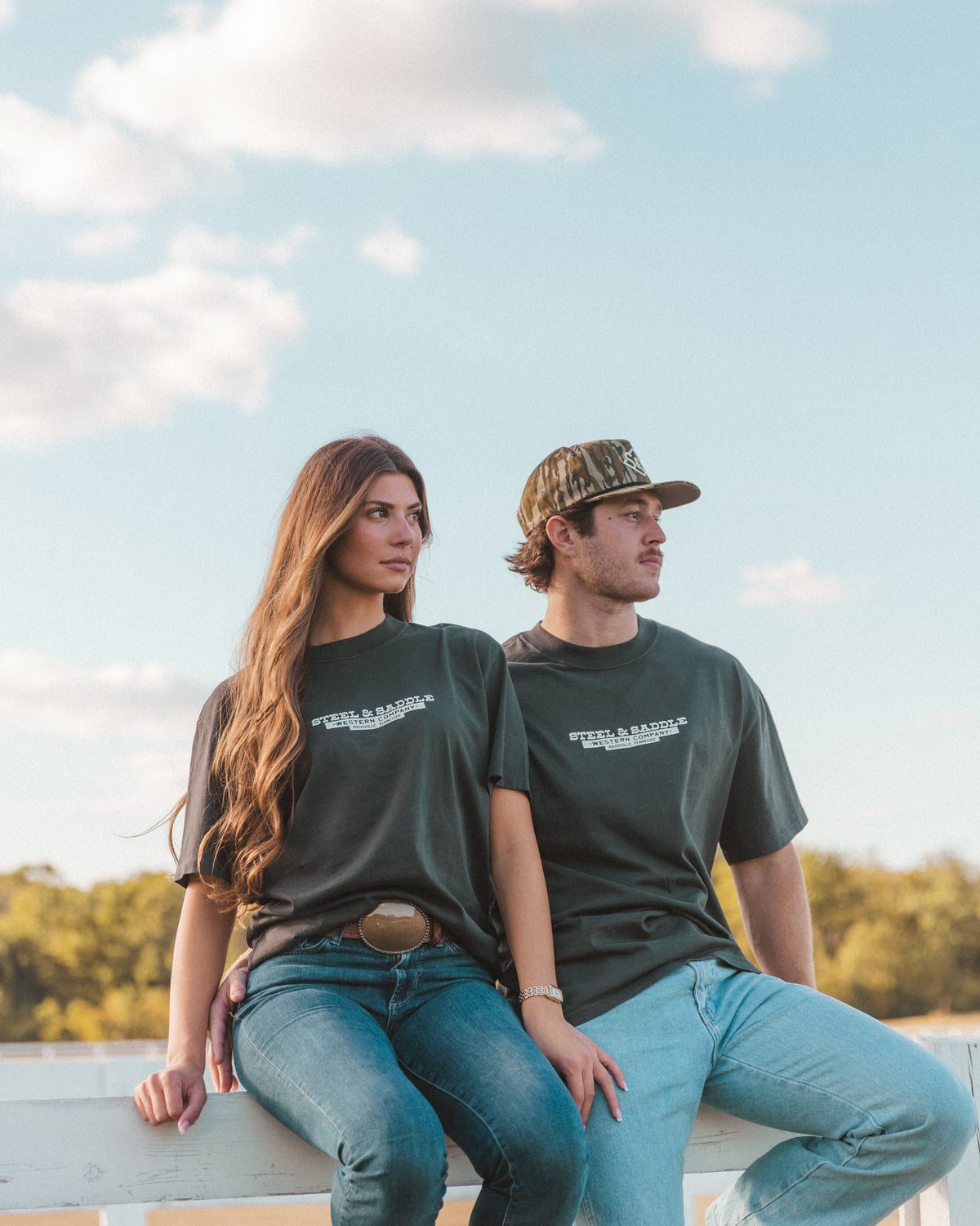 Couple in Steel & Saddle tees sitting together on a white ranch fence