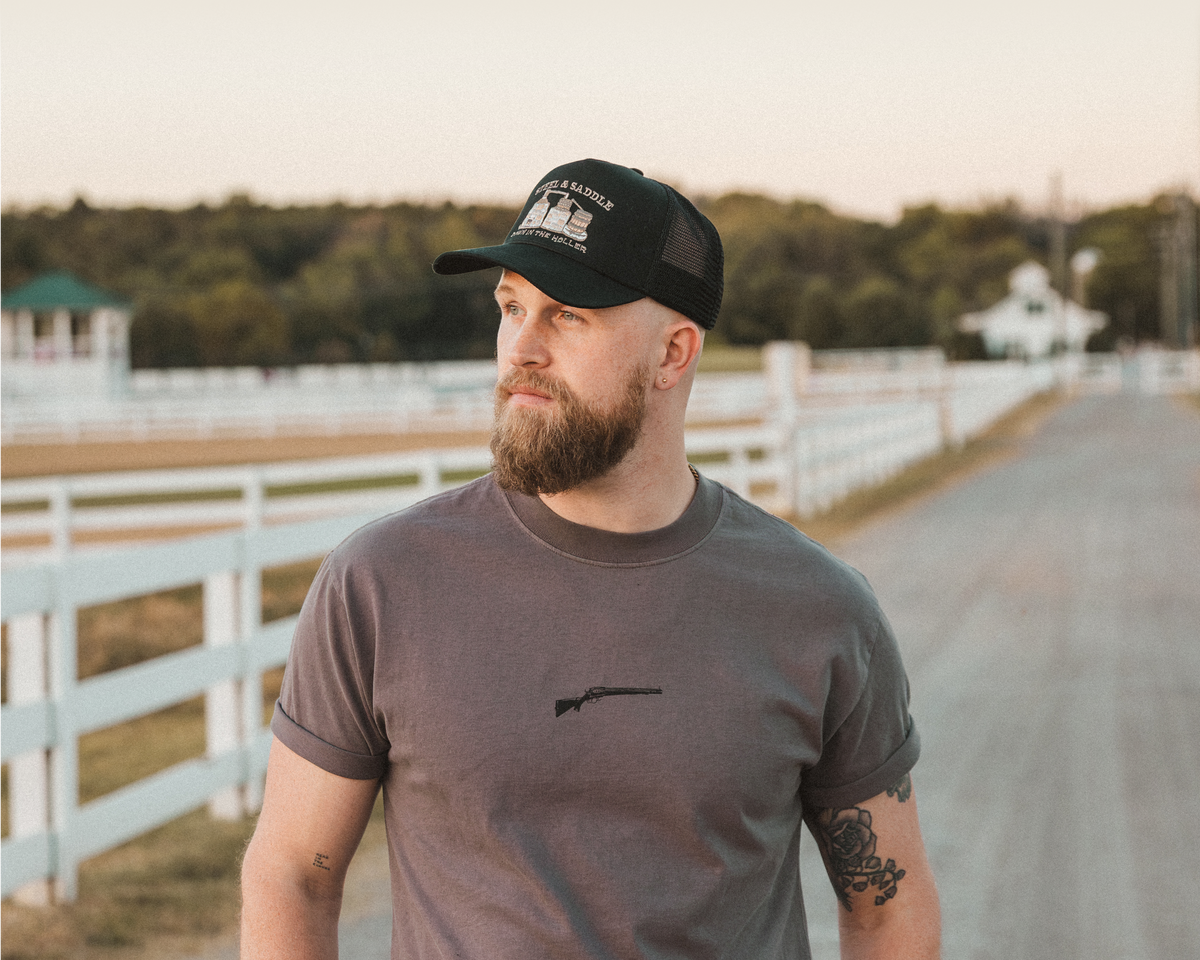 A man wearing a dark gray t-shirt with a small black rifle graphic on the chest and a black trucker hat, standing on a country road with white fences in the background.