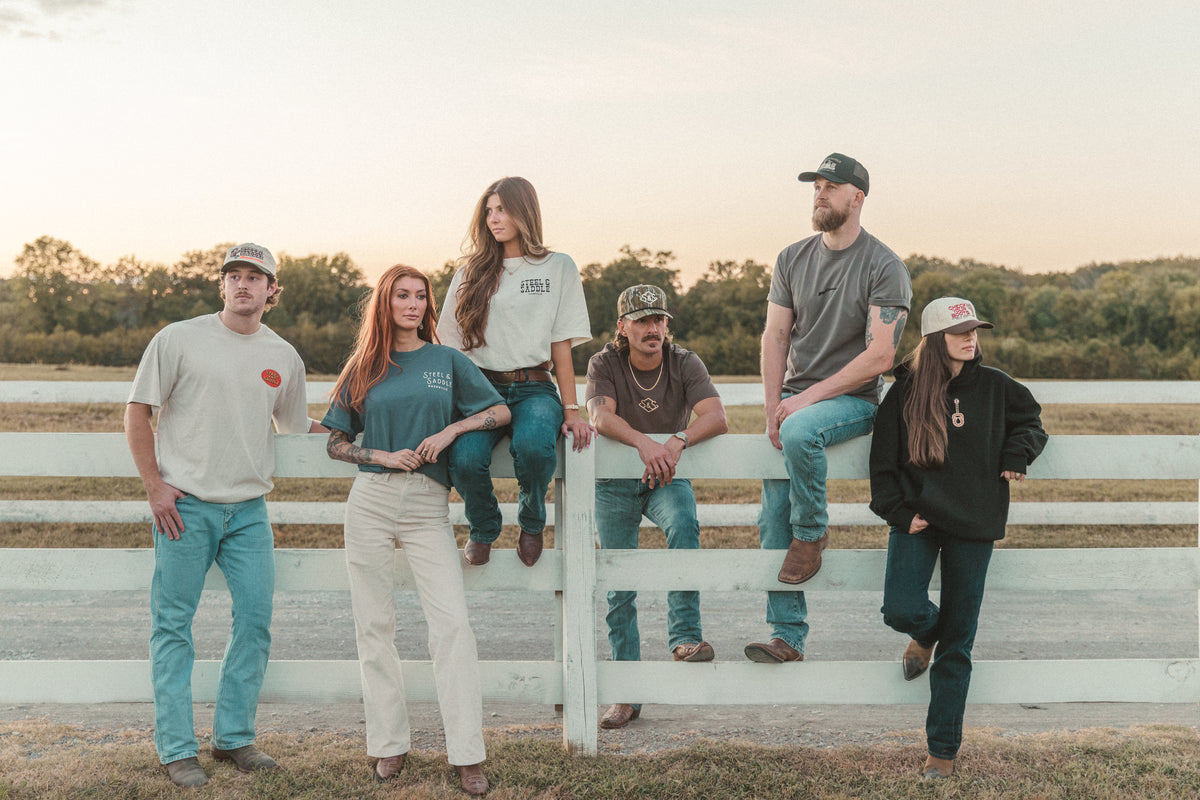 Six friends in Steel & Saddle tees and caps sitting on a white ranch fence at sunset
