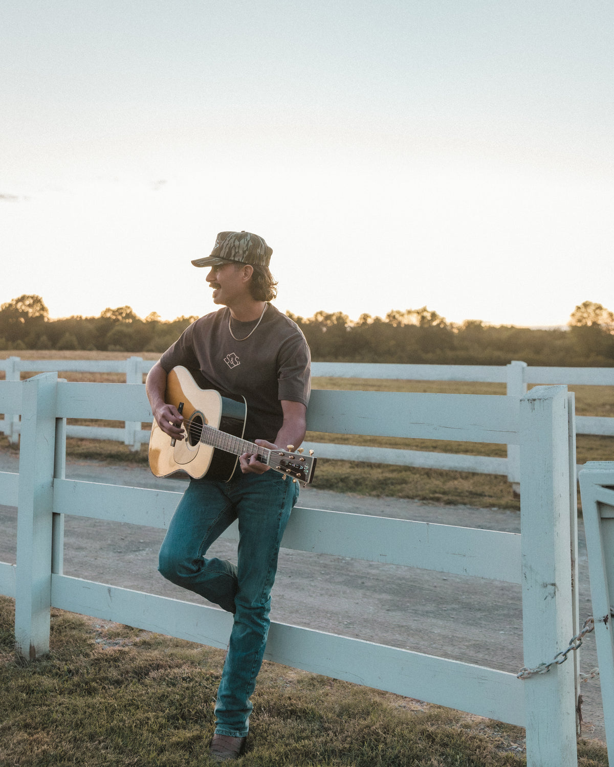 Man in a camo cap leaning on a ranch fence and playing acoustic guitar