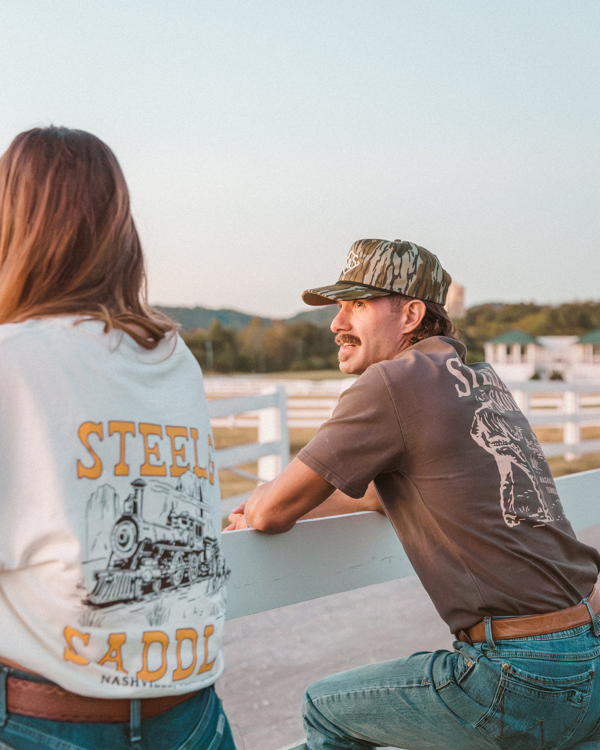 Woman showing the Holdup Tee train robbery back print beside a man in a camo cap at a white fence