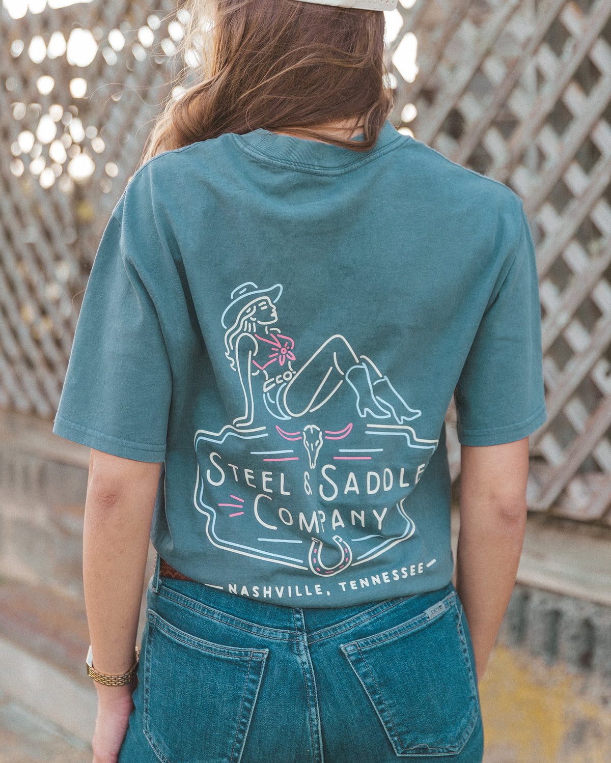 Music City Neon Tee in faded indigo worn outdoors, back print of a neon cowgirl on a Steel & Saddle Company sign